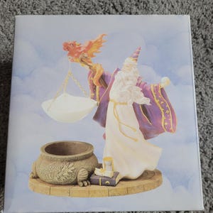 May include: A white ceramic wax warmer with a purple and gold wizard figurine holding a red and orange dragon. The wizard is standing on a base with a book, an hourglass, and a cauldron.