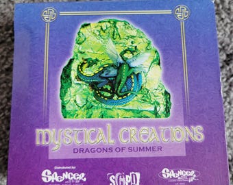 Könnte beinhalten: Quadratische Schachtel mit lila und blauem Hintergrund mit einem grünen und blauen Drachen auf einem grünen Felsen. Der Text "Mystical Creations Dragons of Summer" ist in goldfarbenen Buchstaben dargestellt. Die Schachtel hat goldfarbene Verzierungen und keltische Knotenmuster.