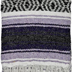 May include: A woven blanket with a striped pattern in shades of purple, white, and black. The blanket has a fringe along the bottom edge. The design includes a central band of black geometric shapes.