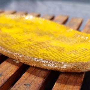 May include: A handmade, oblong ceramic dish with a bright yellow interior and a textured surface. The dish has a brown, unglazed rim. It is set against a wooden slatted background. This decorative piece is suitable for serving or display.