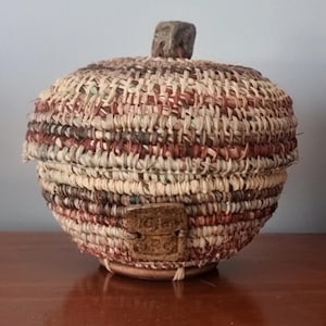 May include: A round woven basket with a lid, made from natural and reddish-brown fibres. The lid has a small, square wooden knob. A small, rectangular wooden tag is attached to the side of the basket.