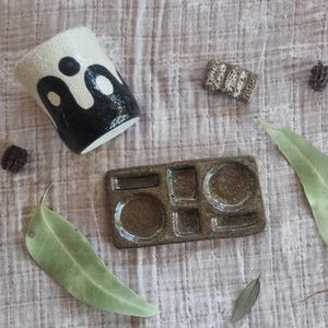 May include: A ceramic cup with a black and white abstract design, a small rectangular object, and a ceramic tray with various compartments. The items are arranged on a textured, neutral-toned surface, with leaves and small pine cones.