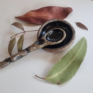 May include: A handmade ceramic spoon and a small matching bowl with a textured, speckled tan exterior and a glossy, dark black interior finish. The spoon features a rustic, uneven handle design, and both pieces are arranged with natural leaves.