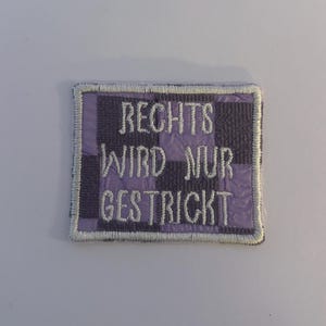 Könnte beinhalten: Ein rechteckiger Aufnäher mit kariertem lila und grauem Hintergrund und weiß gesticktem Text, der "RECHTS WIRD NUR GESTRICKT" lautet. Der Aufnäher hat einen weißen Rand.