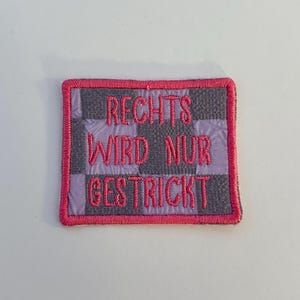 Könnte beinhalten: Gestickter Aufnäher mit dem deutschen Text "RECHTS WIRD NUR GESTRICKT" in Rosa. Der Aufnäher hat ein grau-lila kariertes Muster mit einem rosa Rand. Der Aufnäher ist rechteckig.