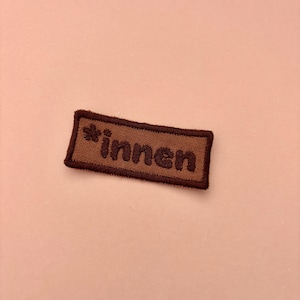 Könnte beinhalten: Ein rechteckiger, bestickter Aufnäher mit dunkelbraunem Rand und hellbrauner Mitte. Der Text "innen" ist in dunkelbraunem Garn gestickt und wird auf der linken Seite von einem kleinen, dunkelbraun gestickten Blumensymbol begleitet.