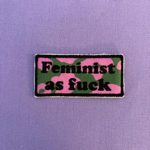Könnte beinhalten: Ein rechteckiger Aufnäher mit den Worten "Feminist as fuck" in schwarzen Buchstaben. Der Hintergrund des Aufnähers hat ein rosa-grünes Tarnmuster, und der Aufnäher ist schwarz umrandet. Der Aufnäher befindet sich auf einem lila Hintergrund.