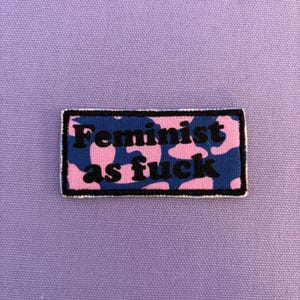 Könnte beinhalten: Ein rechteckiger Aufnäher mit dem Text "Feminist as fuck" in schwarzen Buchstaben. Der Hintergrund des Aufnähers ist blau mit rosa Flecken, und der Rand ist weiß. Der Aufnäher befindet sich auf einer lila Stoffoberfläche.