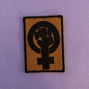 Könnte beinhalten: Rechteckiger Aufnäher mit einem schwarzen Faustsymbol in einem Kreis, über einem Kreuzsymbol, auf beigem Hintergrund. Der Aufnäher hat einen schwarzen Rand und liegt auf einem hellvioletten Stoff.