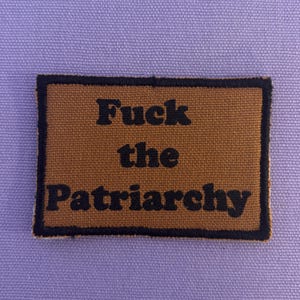 Könnte beinhalten: Ein rechteckiger Aufnäher mit dem Text "Fuck the Patriarchy" in schwarzen Buchstaben auf braunem Grund. Der Aufnäher hat einen schwarzen Rand und ist auf einem lavendelfarbenen Stoff angebracht.