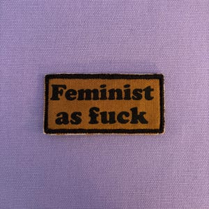 Könnte beinhalten: Ein rechteckiger Aufnäher mit dem Text "Feminist as fuck" in schwarzen Buchstaben auf beigem Hintergrund. Der Aufnäher hat einen schwarzen Rand und befindet sich auf einem hellvioletten Stoffhintergrund.
