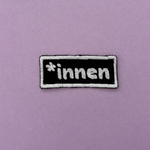 Könnte beinhalten: Ein rechteckiger gestickter Aufnäher mit schwarzem Hintergrund und weißem Rand. Der Text *innen ist in weißen Kleinbuchstaben in der Mitte des Aufnähers gestickt, vorangestellt ist ein kleines weißes sternförmiges Blumensymbol.