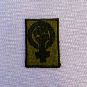 Könnte beinhalten: Ein rechteckiger Aufnäher mit einem schwarzen Faust-in-Kreis-Symbol über einem Kreuzsymbol auf grünem Hintergrund. Der Aufnäher hat einen schwarzen Rand. Das Design ist ein Symbol des Feminismus.