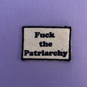 Könnte beinhalten: Ein rechteckiger Aufnäher mit dem Text "Fuck the Patriarchy" in schwarzen Buchstaben auf beigem Hintergrund, umrahmt von einem schwarzen Rand. Der Aufnäher befindet sich auf einem einfarbigen lila Stoff.