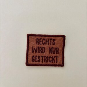 Könnte beinhalten: Ein rechteckiger Stoffaufnäher mit braunem Hintergrund und dunkelbraunem Rand. Der Aufnäher zeigt den dunkelbraun gestickten Text RECHTS WIRD NUR GESTRICKT, der in drei Zeilen mittig innerhalb des rechteckigen Rahmens angeordnet ist.