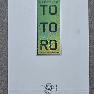 May include: A white card with a green and holographic design that reads "TO TO RO" in large letters. The word "PRESTIGE" is at the top. A small illustration of a character is at the bottom.