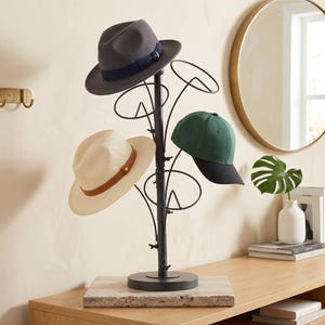 May include: A black metal hat rack with a central pole and multiple circular wire branches for storage. The rack holds a grey fedora with a dark blue band, a cream-colored fedora with a tan leather band, and a green baseball cap with a black brim.
