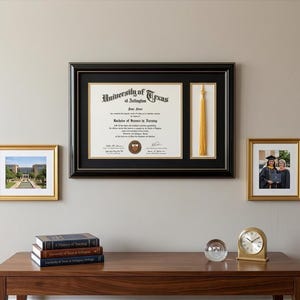 May include: A framed diploma from the University of Texas at Arlington, with a gold tassel, and two framed photos. The diploma is in a black frame with a gold border. The photos are in gold frames. A stack of books and a clock are on a wooden table.