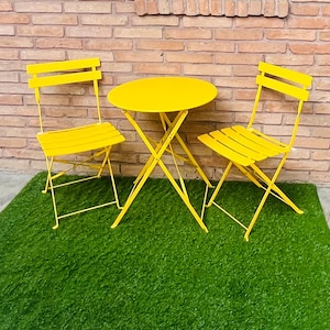 Puede incluir: Un conjunto de bistró amarillo brillante con dos sillas plegables y una mesa redonda. El conjunto es de metal y se encuentra sobre una superficie verde frente a una pared de ladrillo. La mesa y las sillas son de un amarillo vibrante y soleado.
