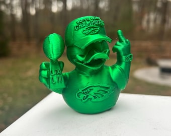 May include: A green duck figurine wearing a baseball cap with the text "SUPER BOWL CHAMPIONS" and an Eagles logo. The duck is holding a trophy and giving the middle finger.