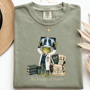 May include: A sage green t-shirt featuring a graphic of a frog dressed in a white lab coat, holding an X-ray, standing next to a stack of books and a tote bag labeled Radiology Is My Superpower. The text We Image at Dawn is printed below.