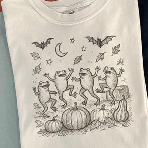 May include: A white t-shirt featuring a whimsical Halloween design. The graphic includes dancing frogs, pumpkins, bats, leaves, a crescent moon, and stars. The shirt is part of a collection of apparel.