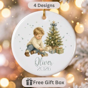 May include: A round white ceramic Christmas ornament featuring a gold hanging string and a printed illustration of a young child playing with a toy car near a decorated Christmas tree with gifts. The text reads 4 Designs, Oliver, and 2026.