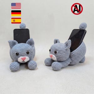 Może przedstawiać: Dwa szare, szydełkowane uchwyty na telefon w kształcie kota. Każdy kot ma okrągłą głowę, spiczaste uszy, biały pyszczek z różowym nosem i czarne oczy. Koty znajdują się w pozycji leżącej, przystosowanej do podtrzymywania czarnego smartfona.