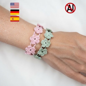 May include: Two crochet bracelets featuring a series of connected five-petaled flower motifs. One bracelet is light pink, and the other is sage green. The floral patterns are linked together in a continuous chain design, creating a delicate accessory.