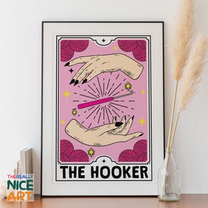 Pode incluir: Um cartão ilustrado rosa e branco com um gancho de crochê rosa e duas mãos segurando-o. O cartão tem o texto "THE HOOKER" na parte inferior.