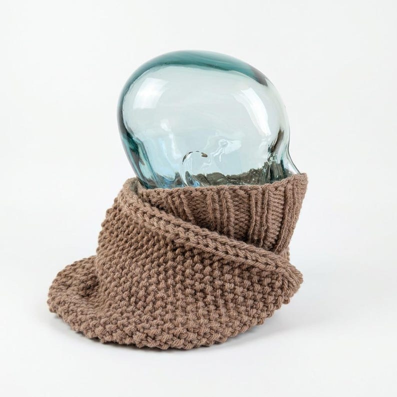 Puede incluir: Una bufanda de cuello tejida de color marr&oacute;n claro con un patr&oacute;n de punto bobo en la secci&oacute;n inferior y un cuello acanalado en la parte superior. La bufanda est&aacute; colocada sobre un expositor liso, transparente y de cristal.