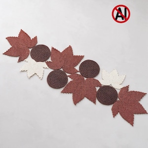 May include: A table runner composed of crocheted elements arranged in a diagonal pattern. The set includes dark brown circular coasters and maple leaf shapes in shades of rust orange and cream, all featuring detailed textured stitching.