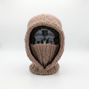 May include: A light brown knitted balaclava featuring a textured stitch pattern on the hood and a ribbed knit design covering the lower face area. The headgear is constructed with a thick, raised border framing the face opening.