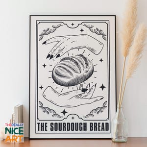 Puede incluir: Lámina artística en blanco y negro que representa una hogaza de pan de masa madre flotando entre dos manos con uñas largas y oscuras. La impresión tiene un borde decorativo y el texto "THE SOURDOUGH BREAD". La obra se exhibe sobre una superficie de madera.
