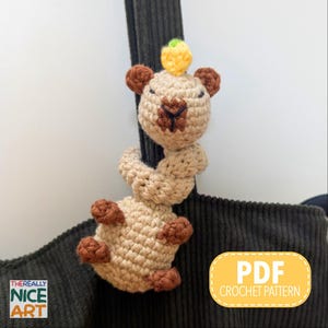 Könnte beinhalten: Ein gehäkeltes Capybara-Spielzeug in Beige, Braun und Gelb, das um einen dunklen Cordriemen gewickelt ist. Das Capybara hat braune Ohren und Füße, eine gelbe Krone und eine dunkelbraune Nase. Das Bild enthält den Text "PDF CROCHET PATTERN".