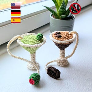 May include: Two crocheted cocktail glass ornaments with cream-colored stems and rims. One glass is filled with light green yarn and topped with a green olive featuring a red center. The other glass contains brown yarn topped with dark brown coffee beans.