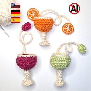 Puede incluir: Tres copas de cóctel tejidas a ganchillo en naranja, magenta y verde claro. Cada copa tiene un tallo y base de color crema, un largo hilo de lana color crema y un pequeño adorno tejido, incluyendo rodajas de naranja y frutas redondas.