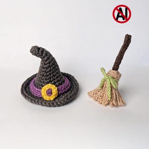 May include: A dark gray crocheted witch hat with a purple band and a yellow circular accent, paired with a small crocheted broom featuring a brown handle and light tan bristles tied with a light green yarn bow, set against a plain background.