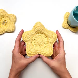 May include: A handmade, yellow crochet coaster in the shape of a starfish. The coaster is held by two hands. Two other coasters are in the background, one holding a blue mug and the other holding two rings. The coasters are a decorative item.