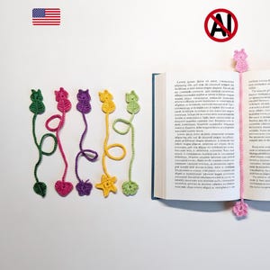 May include: A collection of colorful, handmade crochet bookmarks in various shapes, including cats, hearts, and stars. The bookmarks are in shades of green, pink, purple, yellow, and pink. One bookmark is placed in an open book.