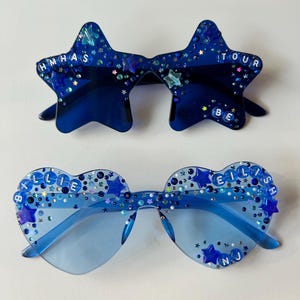 May include: Two pairs of blue sunglasses with star and heart-shaped frames. The star-shaped sunglasses have the text "HMHAS TOUR BE" on them. The heart-shaped sunglasses have the text "BILLIE EILISH" on them. Both pairs of sunglasses are decorated with blue and silver glitter.