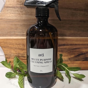 Może przedstawiać: Bursztynowa szklana butelka z rozpylaczem i czarną głowicą. Biała etykieta zawiera napis ard. MULTI-PURPOSE CLEANING SPRAY Tea Tree + Peppermint est. 2024. Świeże zielone liście mięty znajdują się u podstawy butelki po obu stronach.