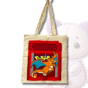 May include: A beige canvas tote bag with a red and gray Pokemon game cartridge design featuring a Charizard Pokemon. The design includes the word "Pok".