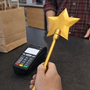 May include: A golden star-shaped wand is held near a black payment terminal. The wand has a long, cylindrical handle and a five-pointed star at the top. A brown paper bag sits on the counter.