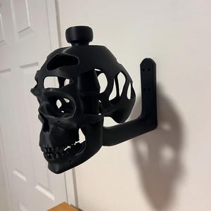 Może przedstawiać: Czarny, 3D-drukowany uchwyt ścienny w kształcie czaszki z wydrążonym wzorem. Czaszka ma mały otwór na górze i wspornik z boku do montażu.