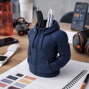 May include: A navy blue pen holder in the shape of a hooded sweatshirt with a zip and pockets. The pen holder contains pens and sits on a wooden desk. Other items on the desk include a water bottle, headphones, and a smartphone.