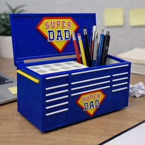 Puede incluir: Un organizador de escritorio azul con forma de caja de herramientas con las palabras "SUPER DAD" en un rombo amarillo y rojo. El organizador está abierto, revelando compartimentos y sosteniendo bolígrafos y lápices. El diseño incluye rayas blancas y detalles amarillos.
