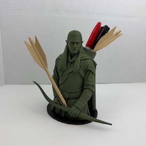 May include: A green figurine of an archer with a bow and arrow. The archer has pointed ears and is wearing a hooded cloak. The figurine holds a bow and arrow, with additional arrows in a quiver. The quiver contains red and black markers.