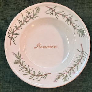 May include: White ceramic plate with a rosemary sprig design. The plate features green rosemary sprigs with purple flowers around the rim and the word "Romarin" in a cursive font in the center. The plate has a light pink rim.