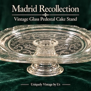 May include: A clear glass pedestal cake stand with a square plate featuring rounded corners and an intricate embossed scrollwork pattern. The stand has a central circular base and includes the text Madrid Recollection.